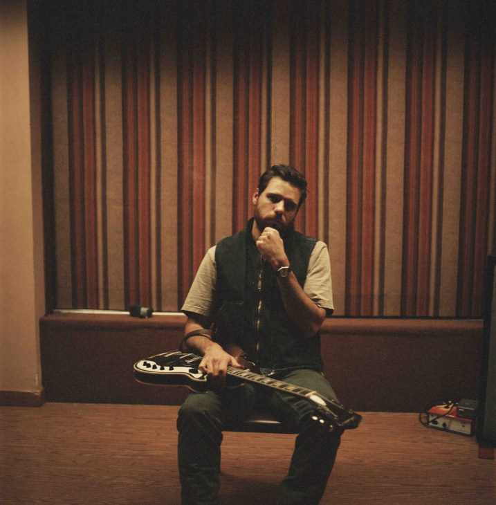 Musician with guitar sitting on stage with red curtains behind him