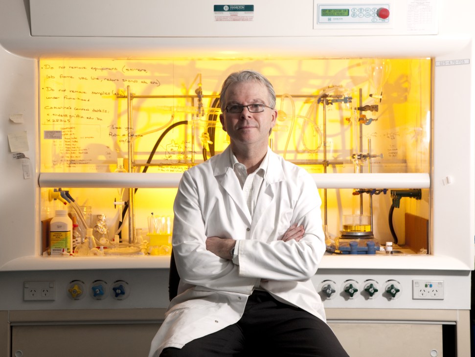 Professor Mark von Itzstein in a white lab coat sits in front of a yellow-lit laboratory fume hood containing scientific equipment and diagrams.