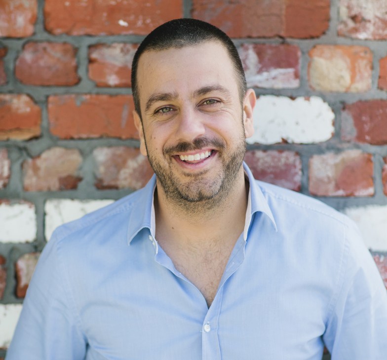 Simon Gloftis, restaurateur and owner of Hellenika, smiles at the camera in front of a brick wall.