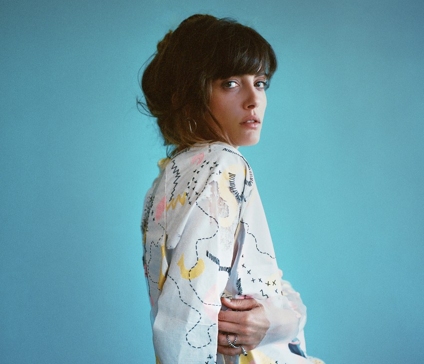 Portrait of Noga Erez wearing a white patterned shirt against a blue background.