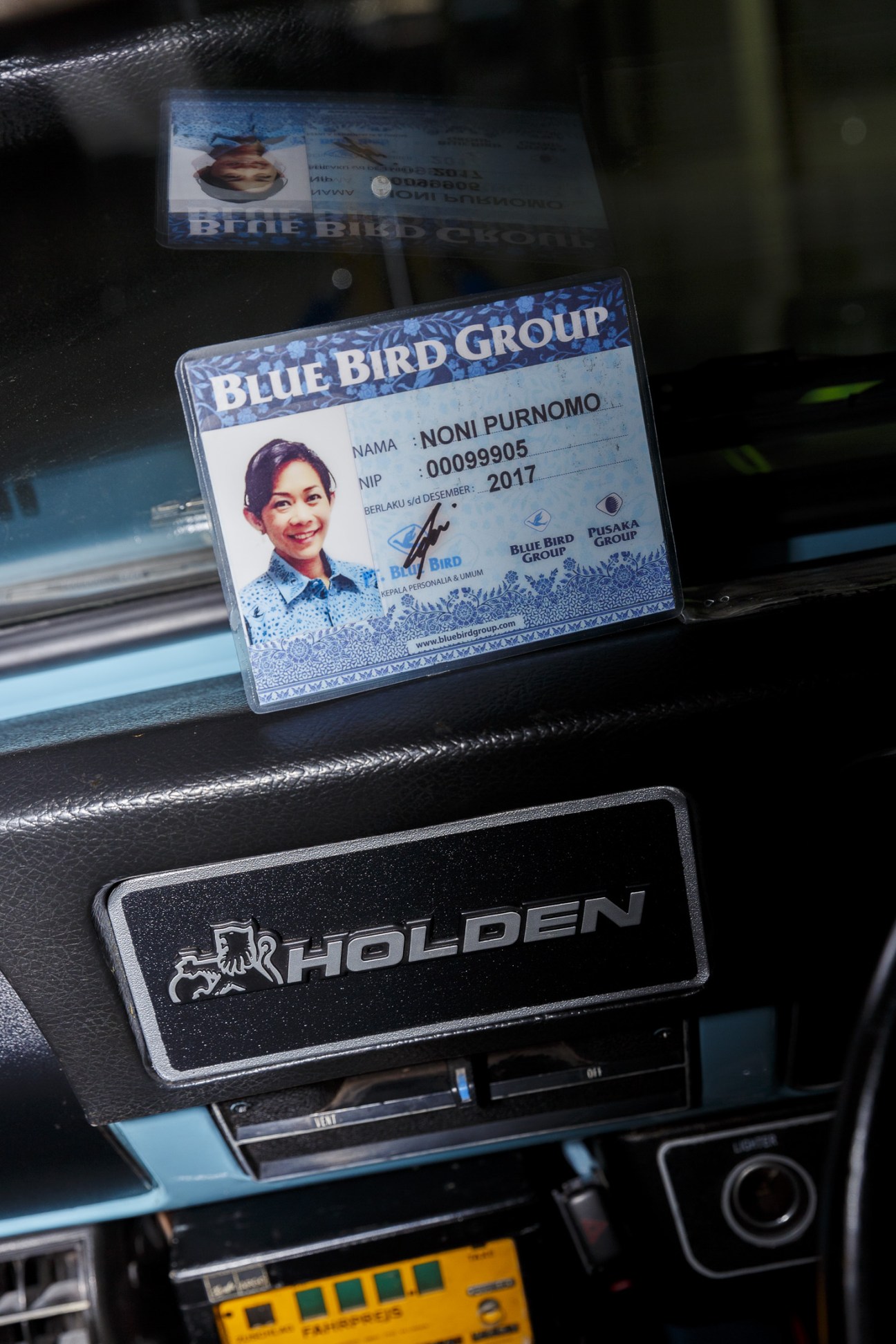 Noni Purnomo's Blue Bird Group taxi driver ID card displayed on a car dashboard.