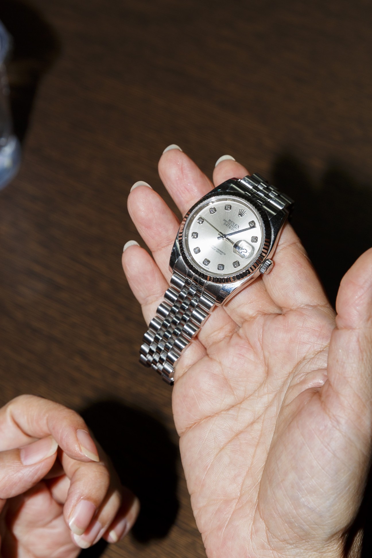 Person's hand holding a silver Rolex watch with diamond hour markers against a brown background.