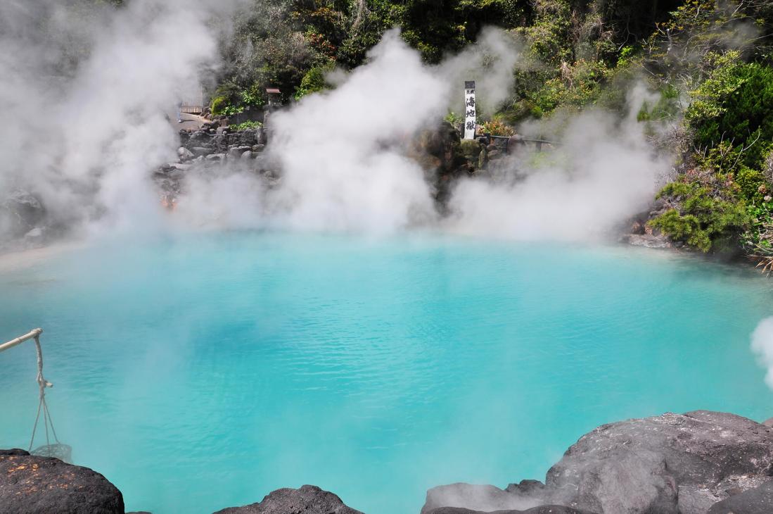 Steaming turquoise hot spring pool surrounded by rocky terrain and lush green vegetation in Beppu, Oita.