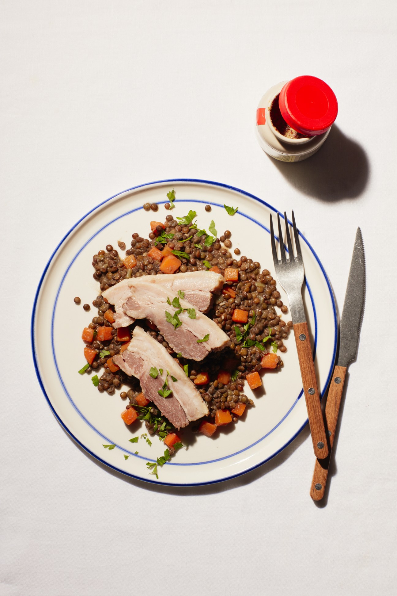 Sliced salted pork served over puy lentils with diced carrots and parsley on a blue-rimmed plate.