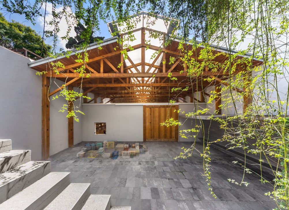 Minimalist courtyard with wooden pergola structure, hanging willow branches, concrete flooring, and white walls.