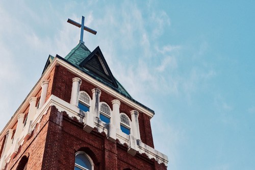 Red-brick church tower with white columns and a cross on top against a blue sky.