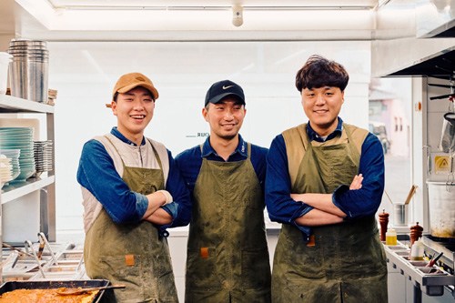 Three people in aprons stand together in a brewery or kitchen space with shelving behind them.
