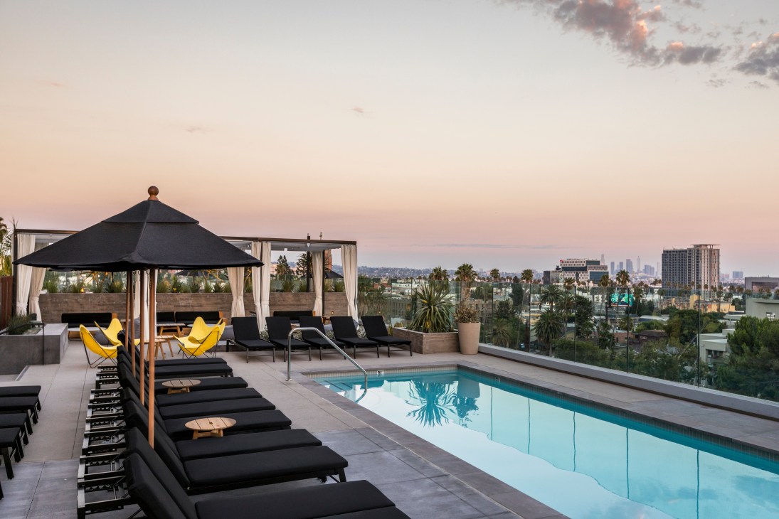 The Everly hotel's fifth-floor pool deck at dusk with sun loungers, black umbrellas, and views toward Los Angeles palm trees and distant cityscape.