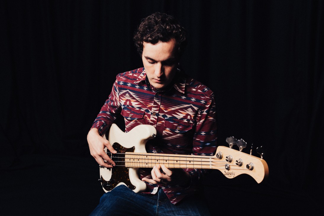 Musician playing white electric bass guitar while wearing patterned maroon shirt against dark background.