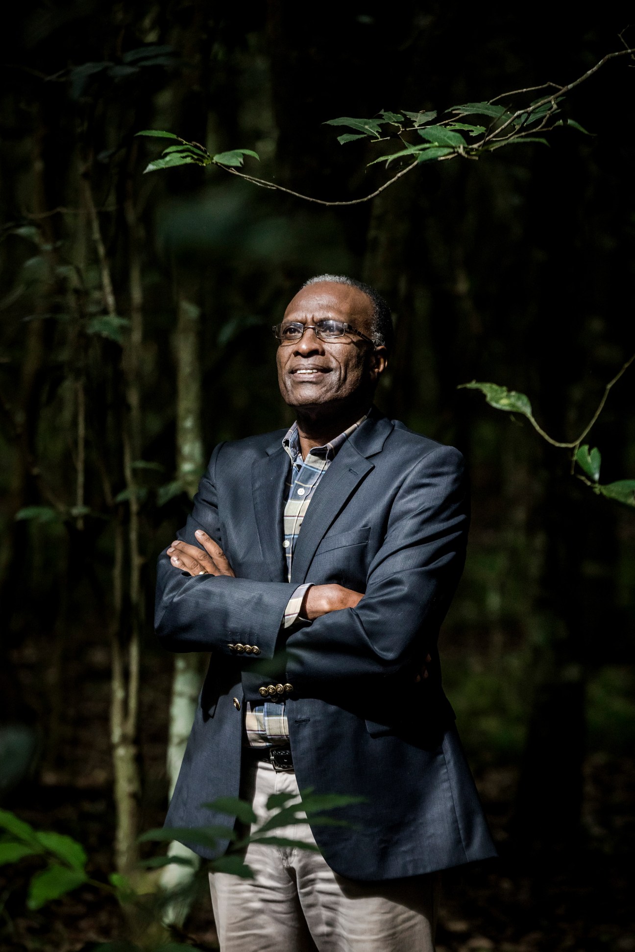 Karanja Njoroge stands with arms crossed in Karura Forest, Nairobi, surrounded by trees and vegetation.