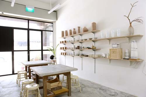 Minimalist retail space with wooden table, woven stools, and shelves displaying ceramic vessels and home goods.