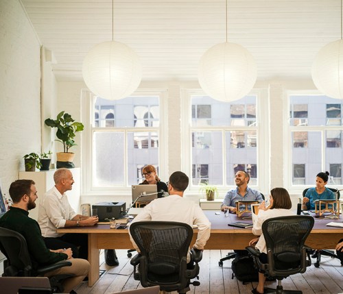 Team meeting at a wooden table in a bright office with high ceilings and large windows overlooking London.