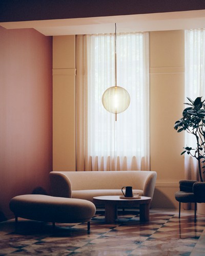 Modern office lobby with curved beige sofa, pendant light, potted plant, and floor-to-ceiling windows with sheer curtains.