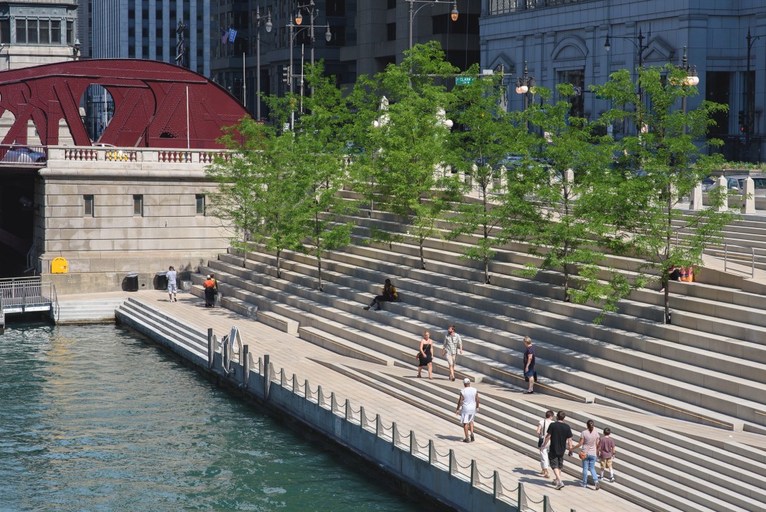 People walking and relaxing on wide stepped plaza beside Chicago River with green trees and modern buildings.