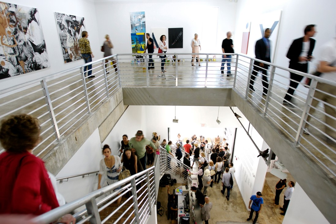 Interior of an art gallery with visitors on multiple levels viewing contemporary artworks on white walls.