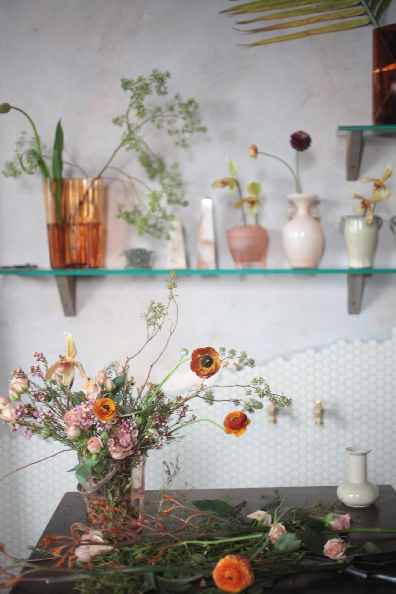 Saipua florist shop display with seasonal flowers in vases on shelves and a vibrant arrangement featuring orange ranunculus and pink carnations.