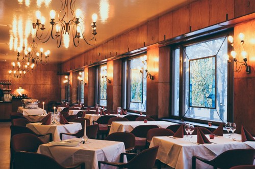 Elegant dining room with wooden tables, white tablecloths, chandeliers and large windows at Meiccu restaurant in Helsinki.