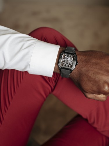 Man wearing white shirt and burgundy trousers displays Cartier Santos de Cartier Skeleton watch on wrist.