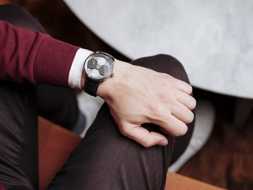 Person wearing burgundy sleeve with white cuff displaying luxury watch with dark dial and leather strap