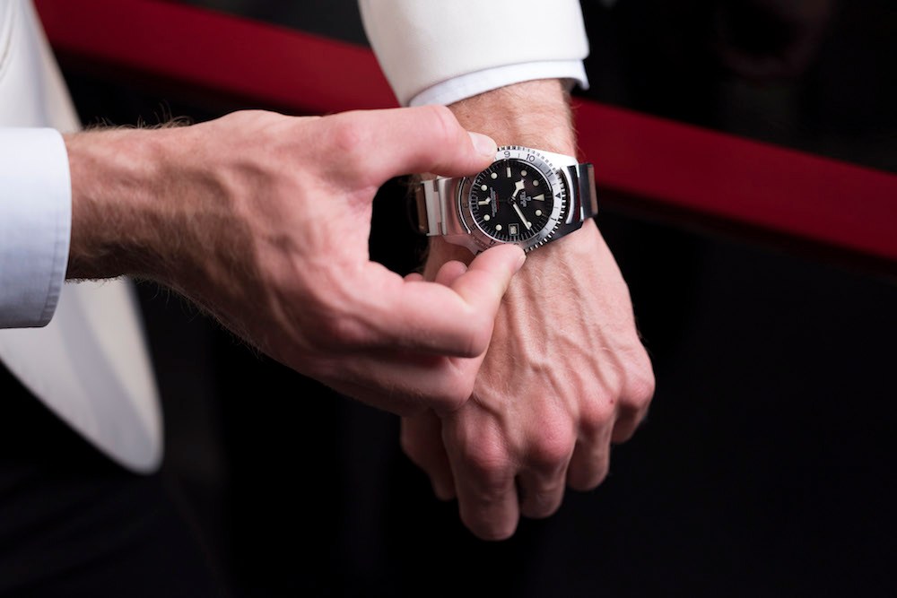 Man's hand wearing Tudor Black Bay P01 watch with black dial and hybrid leather-rubber strap against dark background.