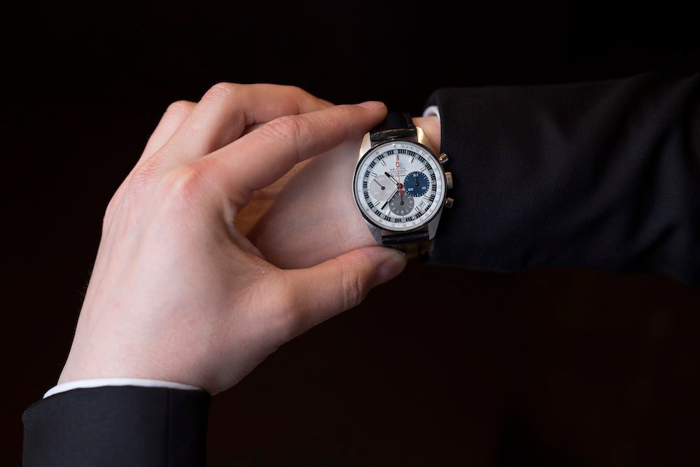 Hand holding Zenith El Primero watch with gold casing and white dial against black background