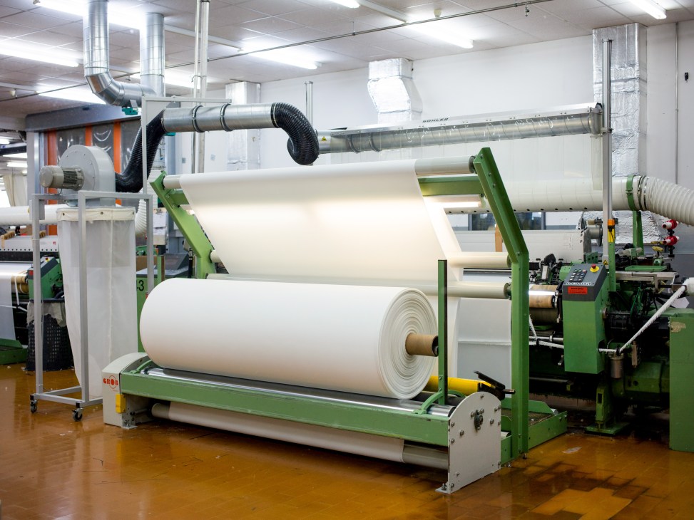 Industrial textile manufacturing facility with large white fabric rolls on green machinery and metal ducting overhead.