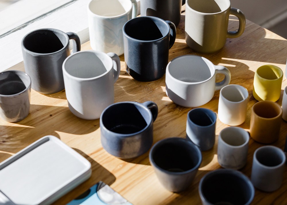 Assorted ceramic mugs in various colors and sizes displayed on a wooden table in natural light.