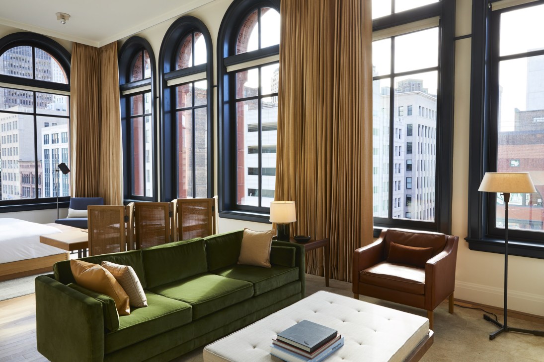 Hotel room at Shinola Hotel with green sofa, brown leather chair, and large windows overlooking Detroit cityscape.