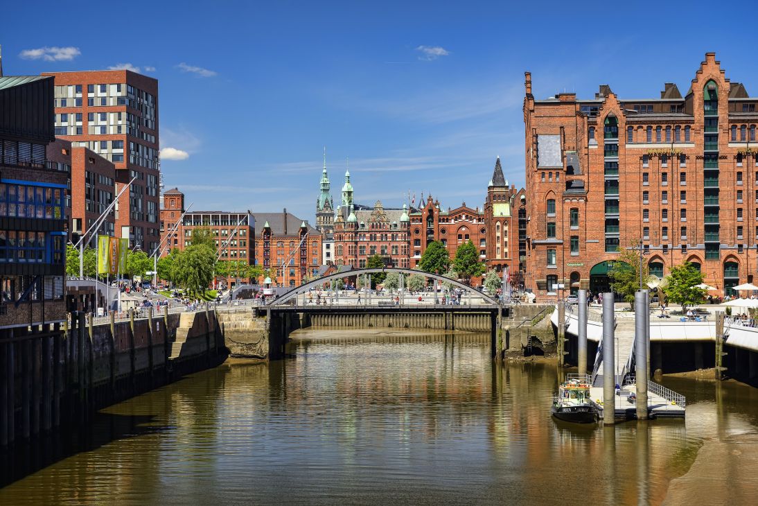 Hamburg's waterfront with red-brick historic buildings, a bridge crossing the canal, and clear blue sky.