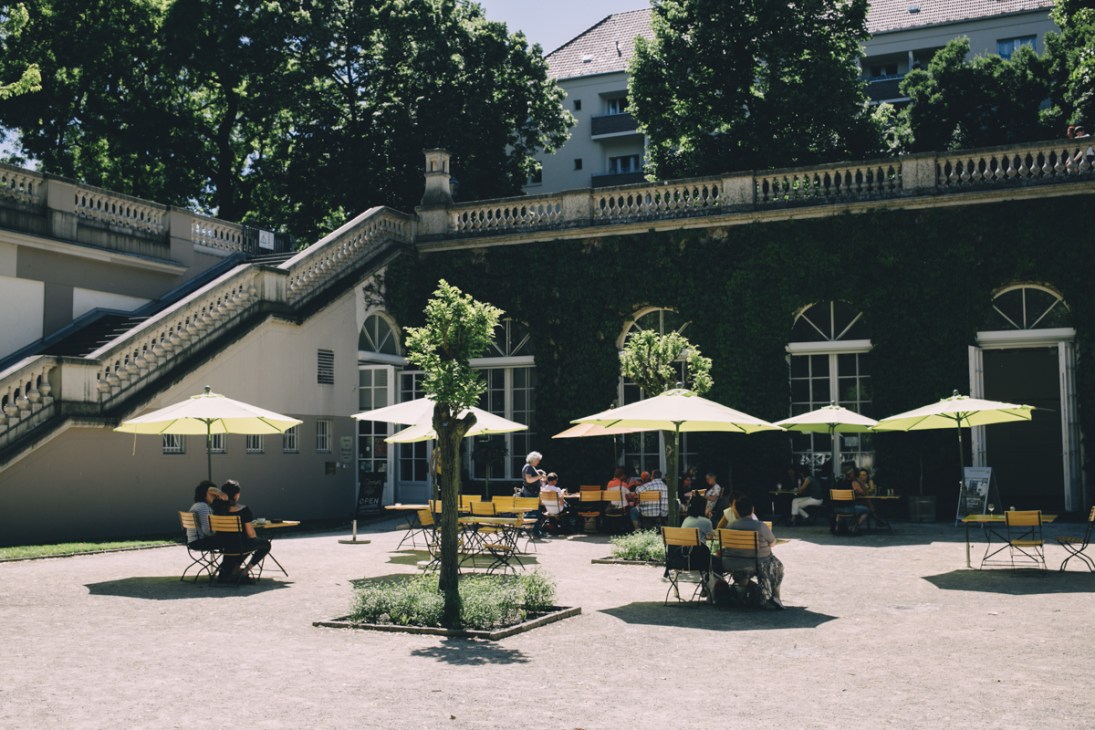 Outdoor café terrace with yellow umbrellas and seating in a neo-baroque park surrounded by ivy-covered walls and historic architecture.