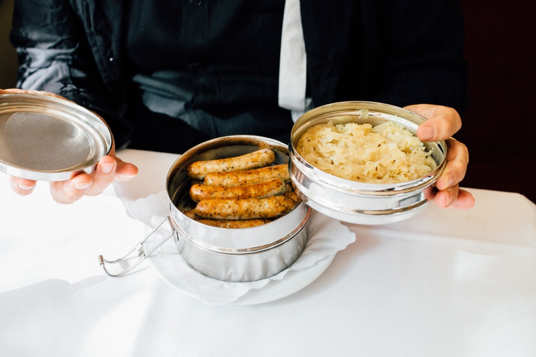 Hands holding a metal lunchbox containing sausages with sauerkraut and mustard.