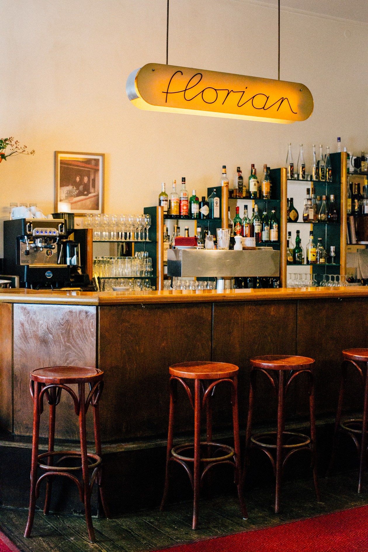 Florian restaurant bar with illuminated sign, wooden counter, bentwood stools, and shelves of bottles.