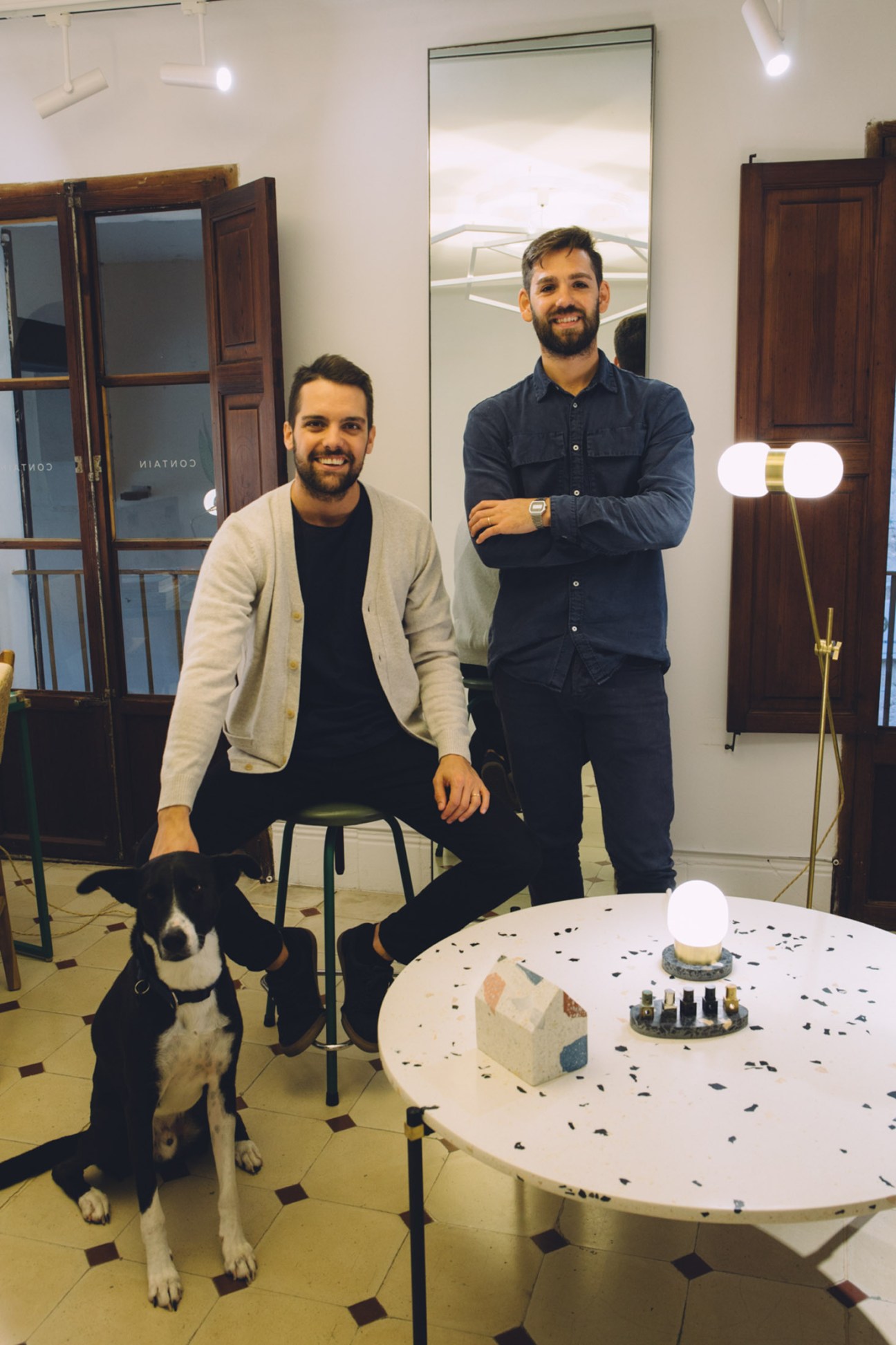 Two men and a dog pose in a modern studio space with white walls, wooden doors, and display lighting.
