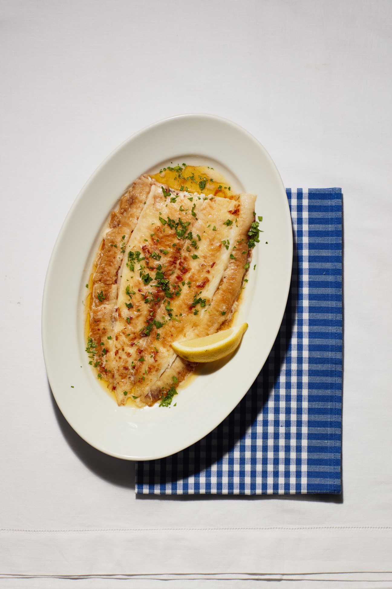 Sole meunière served on a white plate with brown butter, parsley, and lemon wedge on a blue gingham napkin.
