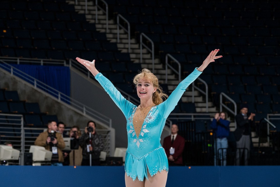 Figure skater in blue sequined dress with arms raised celebrating on ice rink.