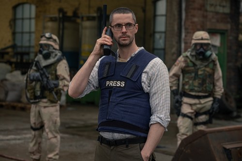 A man wearing a PRESS vest speaks on a radio with armed soldiers in the background.