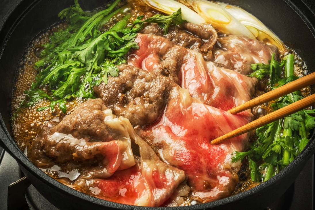 Sukiyaki hot pot with sliced beef, vegetables and noodles in broth with chopsticks.