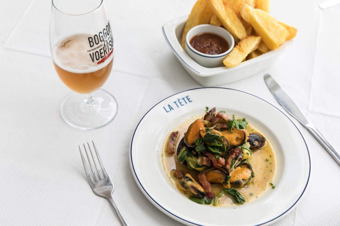 La Tete restaurant dish with seafood, sauce, and garnish alongside a Boggol beer and fries on white table.