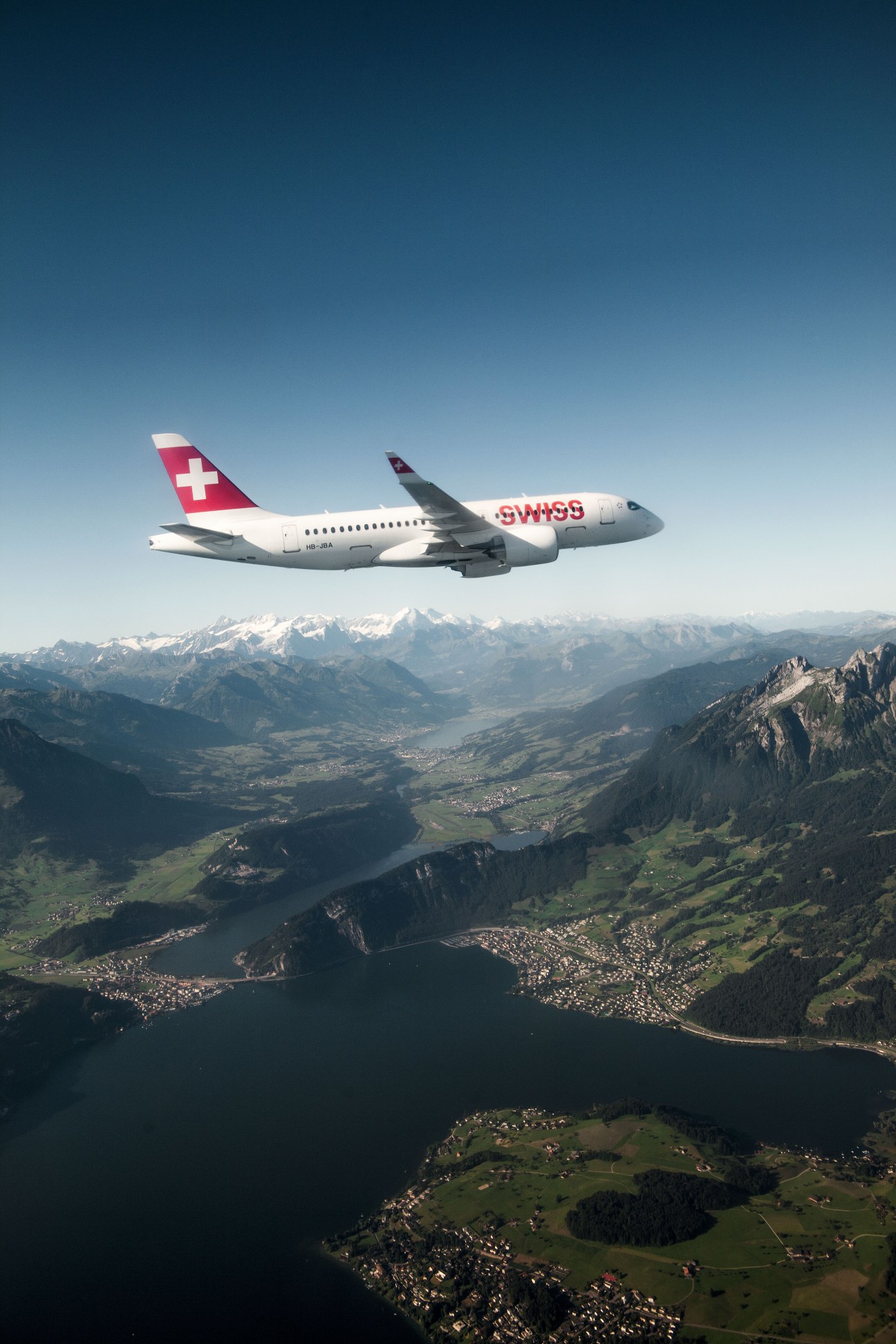 Swiss International Air Lines Bombardier C Series aircraft in flight over Alpine mountains and lake.
