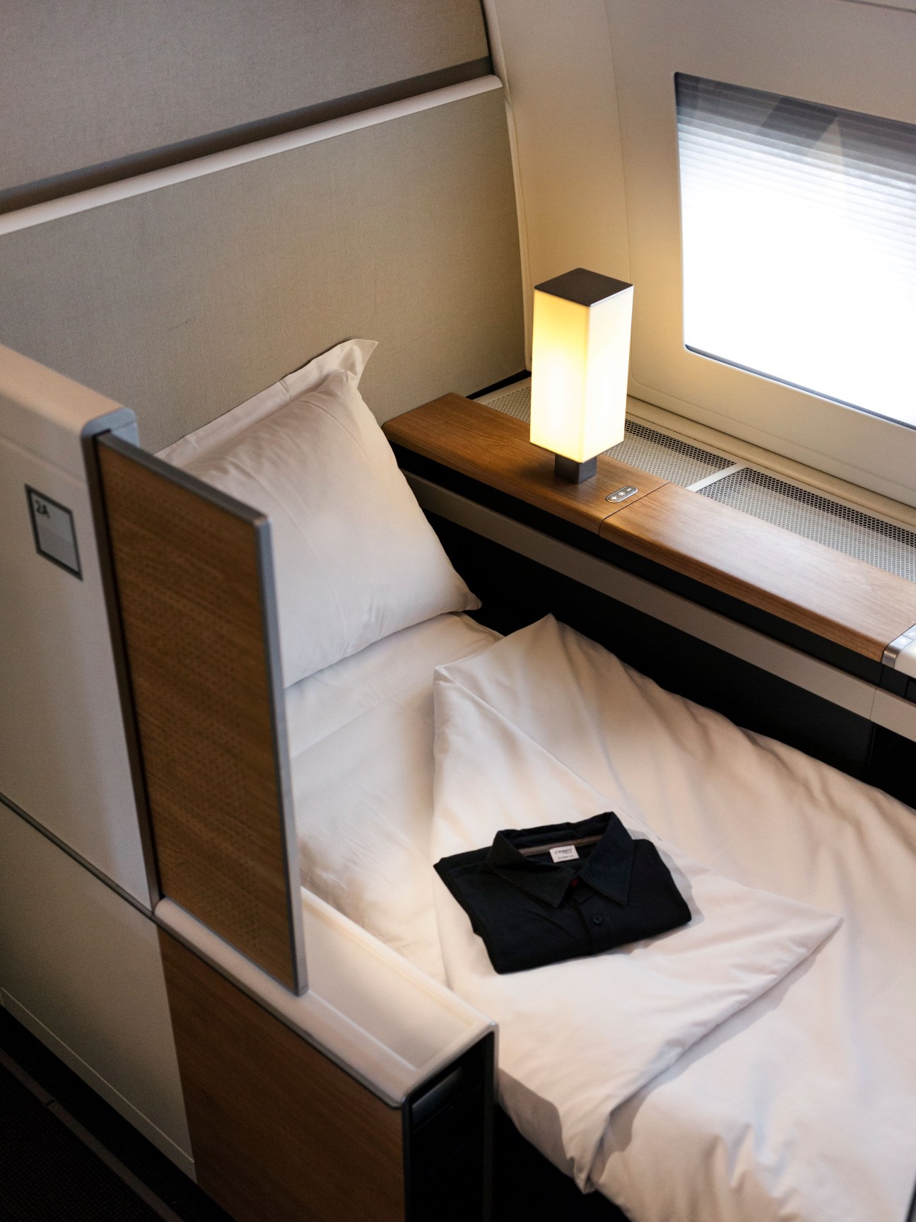 Swiss First Class aircraft cabin bed with white linens, pillow, and illuminated bedside lamp on wooden shelf