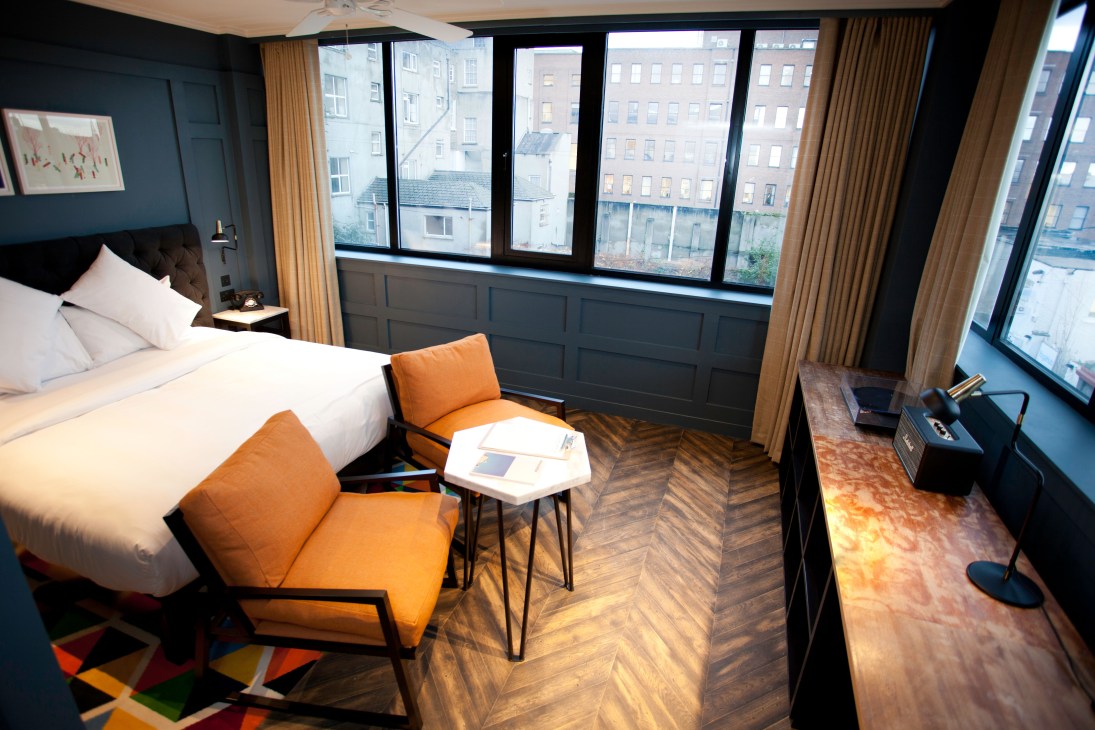 Modern hotel bedroom at The Dean Dublin with orange chairs, white bed, and city views through large windows.