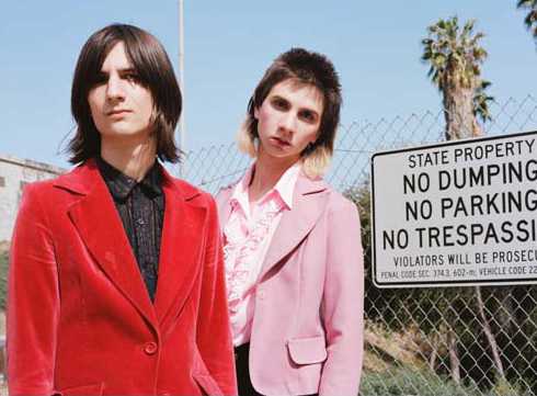 Two young men in colourful blazers pose outdoors against a chain-link fence and palm tree.
