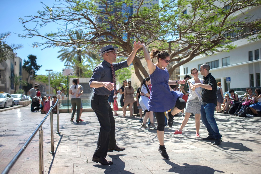 A swing-dance troupe performs in a sunny Tel Aviv street lined with trees and buildings.