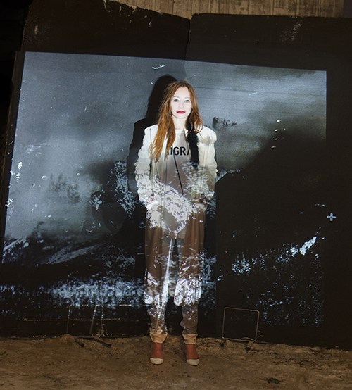 Tori Amos stands against a dark mountainous backdrop wearing a black blazer and brown pants.
