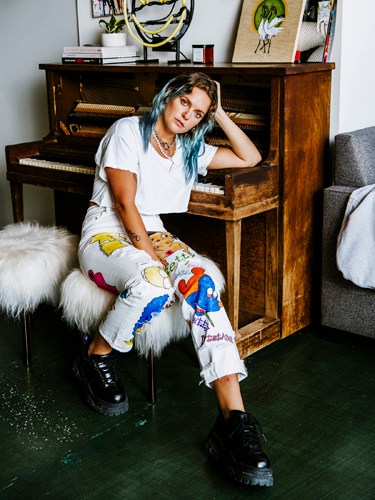 Swedish popstar Tove Lo sits on a stool in front of a wooden piano, wearing a white t-shirt and colorful patterned pants.
