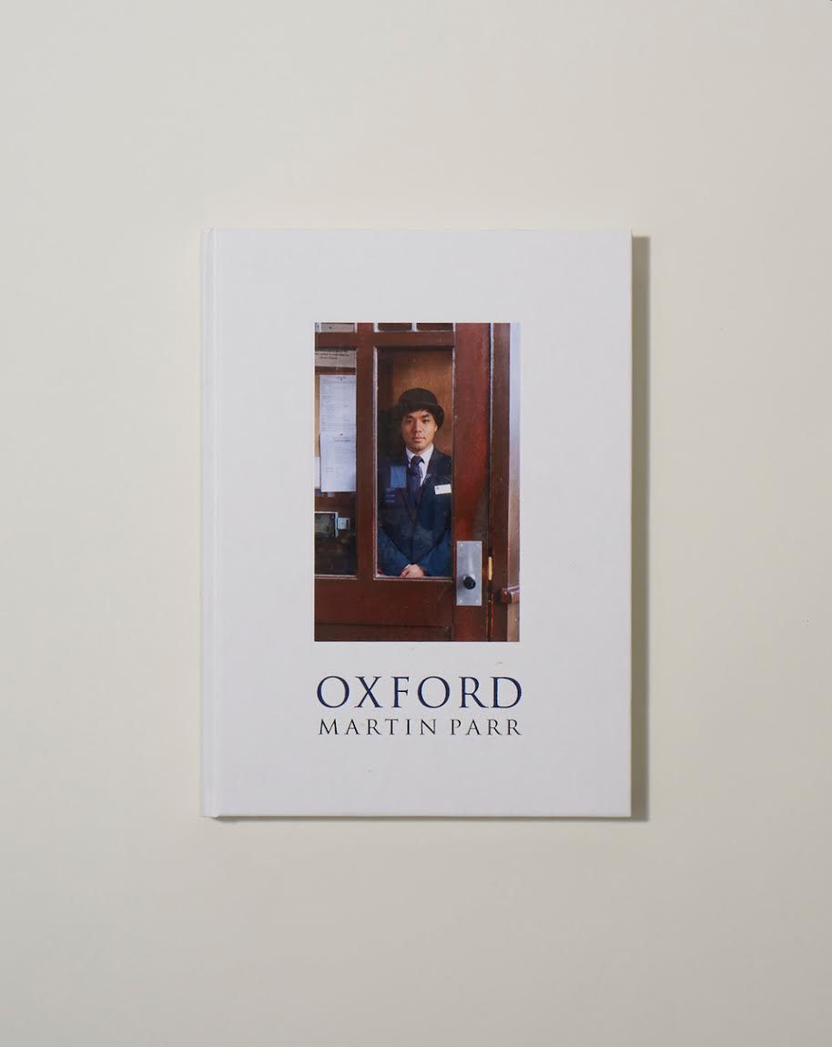 Book cover of Oxford by Martin Parr showing a person in dark clothing looking out from a red wooden doorway or booth.