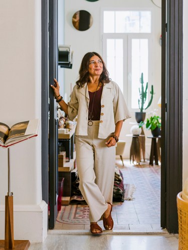 Woman in cream-colored suit standing in a bright hallway with plants and home decor visible.