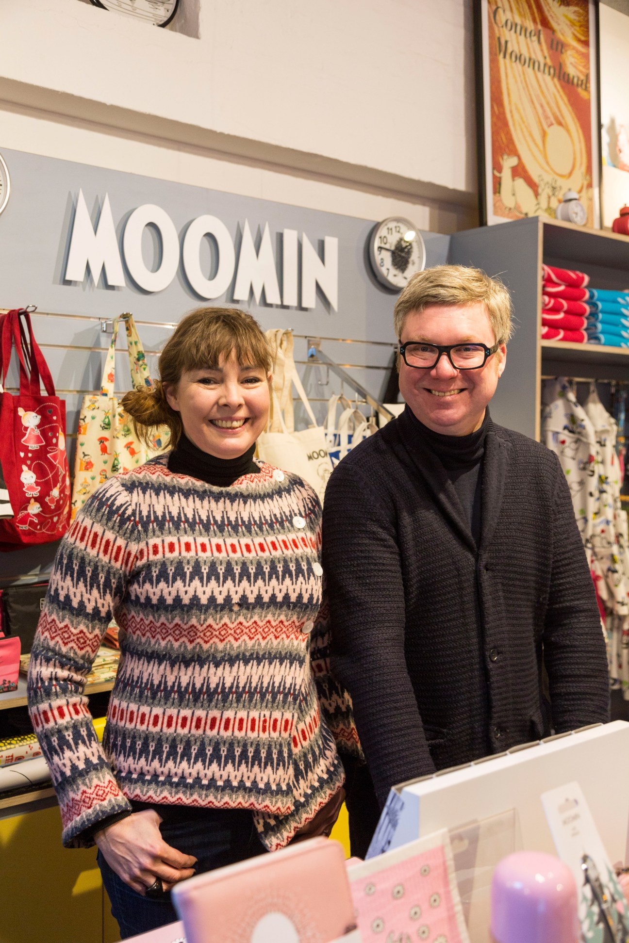 Two people smiling at the Moomin Shop counter in Camden, London, with Moomin branding and colorful products visible behind them.