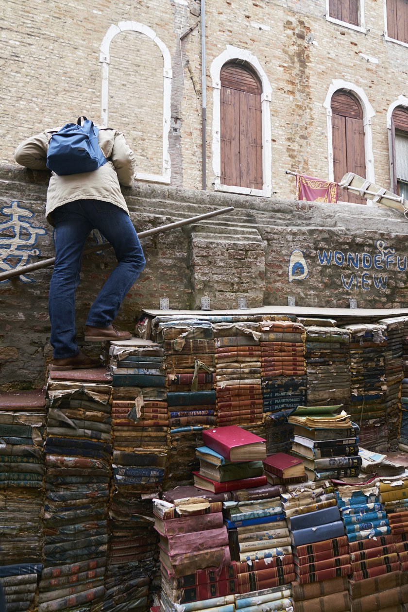 Person climbing stacked vintage books outside a stone building with arched doorways.