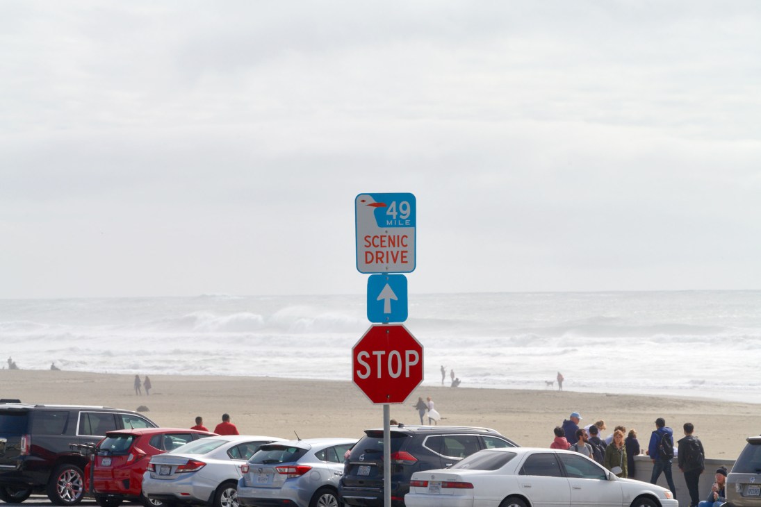 A 49 Mile Scenic Drive sign stacked above a directional arrow and stop sign overlooking a beach with parked cars and visitors.